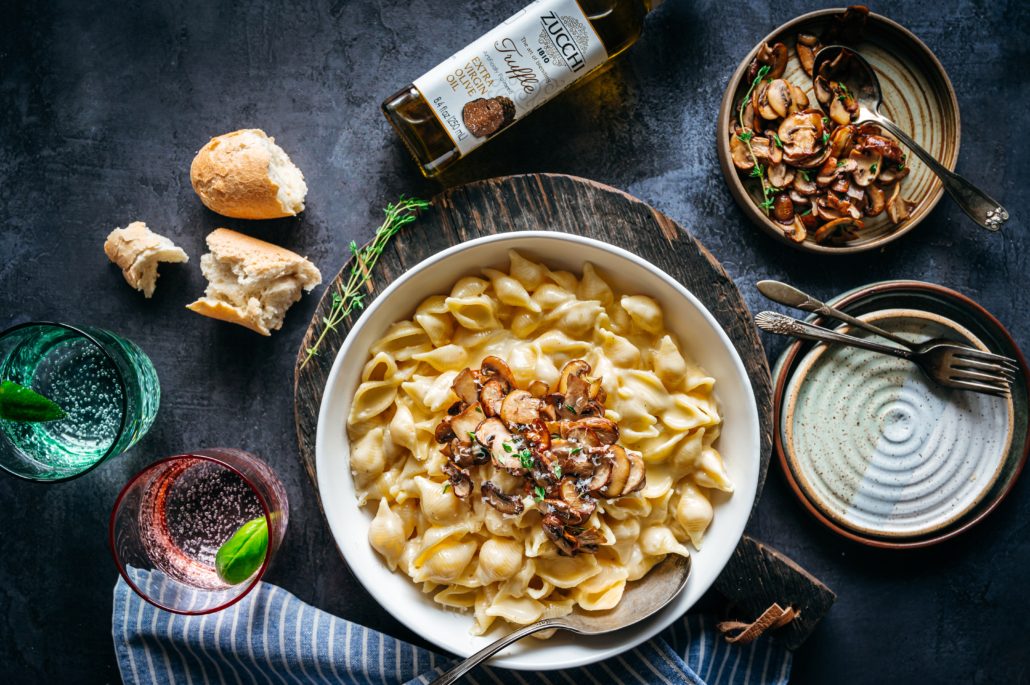 There a dark, bluish grey background with a table setting of food and drink. In the middle is a white bowl filled with golden macaroni and cheese, topped with fresh mushrooms in herbs. Around the bowl are glasses of water, two smaller empty plates with forks, a bowl of cooked mushrooms, torn bread, and a bottle of Zucchi Truffle-Flavored Extra Virgin Olive Oil.