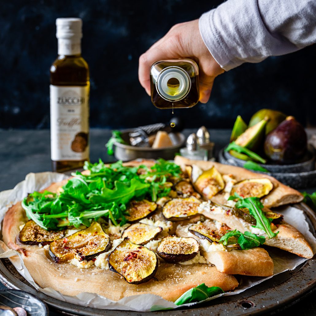 There is a pizza topped with fresh sliced figs, arugula, and cheese. A hand is pouring olive oil from a bottle at the top. In the background there is a bottle of Zucchi truffle-flavored extra virgin olive oil. The backdrop is black and the surface and a bluish grey.