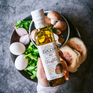 A hand is holding a bottle of Zucchi Truffle-Flavored Extra Virgin Olive Oil. In the background, there is a blue-grey surface with a plate that has two boiled eggs, english muffins, raw mushrooms, and arugula.