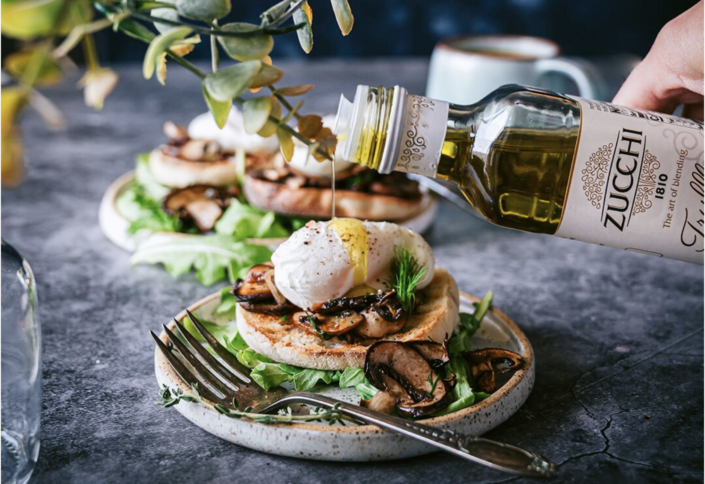 There is a bluish-grey surface with a plate holding an egg dish with arugula, half of an english muffin, cooked mushrooms and a poached egg. There is a hand coming in from the right side, pouring a bottle of Zucchi truffle oil. The oil is drizzling down on top of the poached egg.