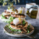 There is a bluish-grey surface with a plate holding an egg dish with arugula, half of an english muffin, cooked mushrooms and a poached egg. There is a hand coming in from the right side, pouring a bottle of Zucchi truffle oil. The oil is drizzling down on top of the poached egg.