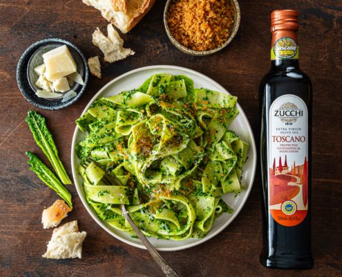 There is a plate of pasta in the center: wide ribbon pappardelle coated in a green kale sauce and topped with toasted breadcrumbs. Surrounding the plate are some of the raw ingredients: a few pieces of torn bread, chunks of Pecorino cheese, a bowl of toasted breadcrumbs, and a bottle of Zucchi 1810's Toscano IGP extra virgin olive oil. Everything sits on a dark coffee brown surface.