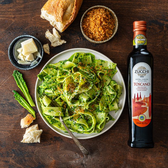 There is a plate of pasta in the center: wide ribbon pappardelle coated in a green kale sauce and topped with toasted breadcrumbs. Surrounding the plate are some of the raw ingredients: a few pieces of torn bread, chunks of Pecorino cheese, a bowl of toasted breadcrumbs, and a bottle of Zucchi 1810's Toscano IGP extra virgin olive oil. Everything sits on a dark coffee brown surface.