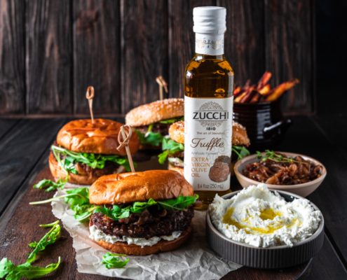 Truffle burger with whipped goat cheese and arugula. In the background we see a bottle of truffle extra virgin olive oil and bowls filled with other ingredients. The surface and background are dark and woody.