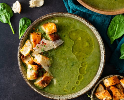 We see a bowl of a green purred soup. on top there are large croutons and thin flakes of cheese, and a drizzle of olive oil. The background is black and moody. The bowl of soup is surrounded by other ingredients including a piece of french bread, a few spinach leaves, and a small bowl of croutons.