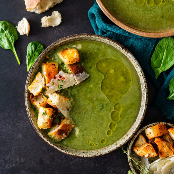 We see a bowl of a green purred soup. on top there are large croutons and thin flakes of cheese, and a drizzle of olive oil. The background is black and moody. The bowl of soup is surrounded by other ingredients including a piece of french bread, a few spinach leaves, and a small bowl of croutons.