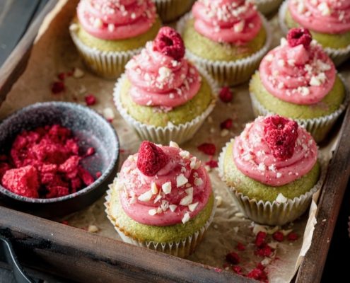 A wooden tray holds several green cupcakes topped with pink frosting and dried raspberries. The cupcakes are garnished with chopped nuts.
