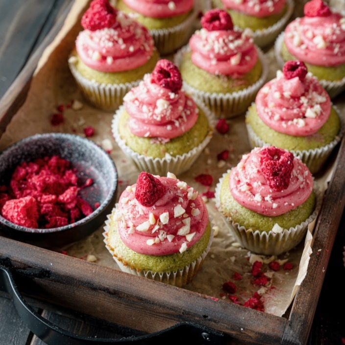 A wooden tray holds several green cupcakes topped with pink frosting and dried raspberries. The cupcakes are garnished with chopped nuts.