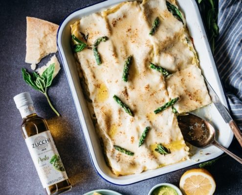 A white rectangular baking tray holds a finished lasagne. The top is creamy and white and studded with green asparagus tips. We see a bottle of olive oil with the words "pesto" on it laying off to the right. The background is a slate blue.