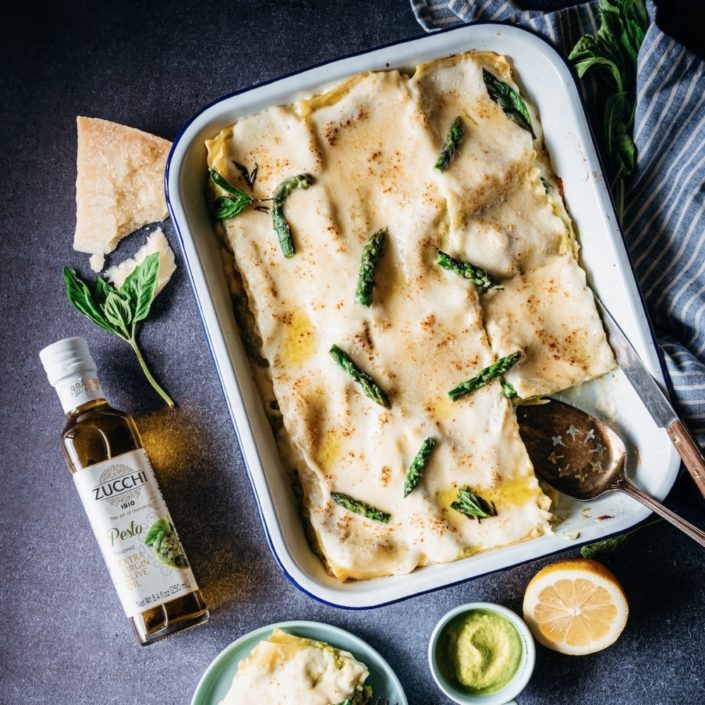 A white rectangular baking tray holds a finished lasagne. The top is creamy and white and studded with green asparagus tips. We see a bottle of olive oil with the words "pesto" on it laying off to the right. The background is a slate blue.