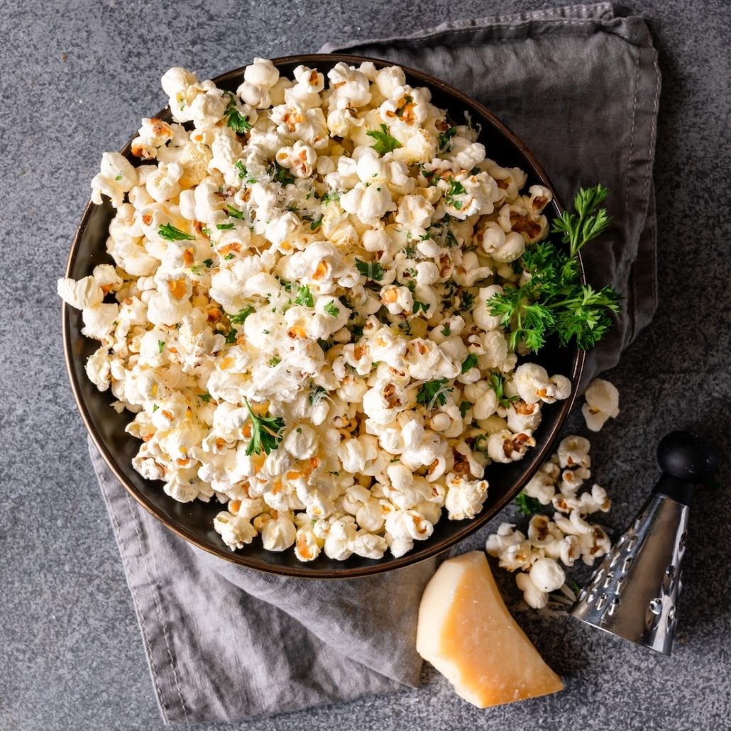 A metal bowl holds freshly popped popcorn garnished with chopped, green parsley. There is a sprig of parsley off to the right. We also see a small metal cheese grater and block of parmesan cheese in the bottom right hand corner. The background is speckled gray.