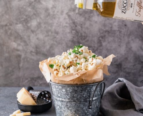 A gray metal pail lined with brown parchment paper is filled with popped popcorn. Above the pail we see a bottle of truffle olive oil drizzling olive oil on top of the popcorn. The back ground is gray and soft.