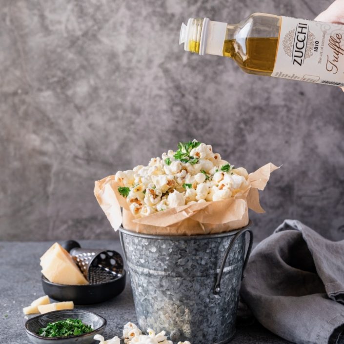 A gray metal pail lined with brown parchment paper is filled with popped popcorn. Above the pail we see a bottle of truffle olive oil drizzling olive oil on top of the popcorn. The back ground is gray and soft.